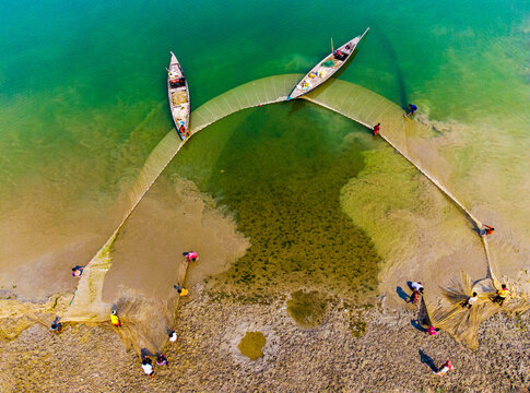 Sariakandi, Bangladesh - 27 May 2024: Aerial view of fishermen casting a wide net in the shallows, the turquoise water contrasting against the golden sand and boats.