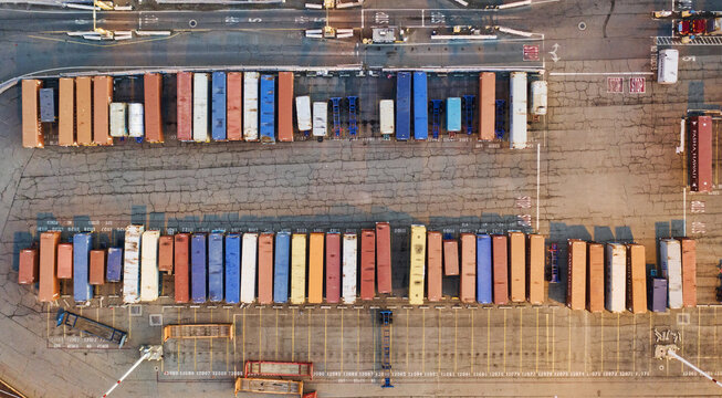 Oakland, United States - 12 May 2020: Aerial view of rows of stacked shipping containers casting long shadows across the cracked asphalt of a bustling port yard.