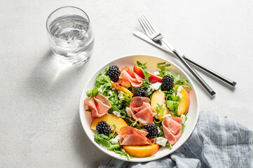 Fresh salad with peaches, arugula, feta, prosciutto and berries in a plate on a light background