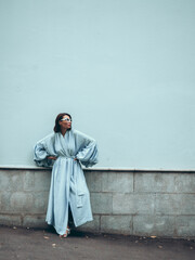 Woman in light blue robe leaning against wall. Stylish woman posing in elegant long robe outdoors. This image of stylish woman in light blue robe is perfect for fashion blogs and websites. © redcat