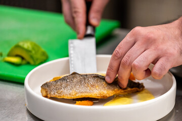 Chef plating crispy fish fillet with yellow sauce and vegetables in professional kitchen. Culinary presentation, gourmet dish preparation, close-up of hands and knife