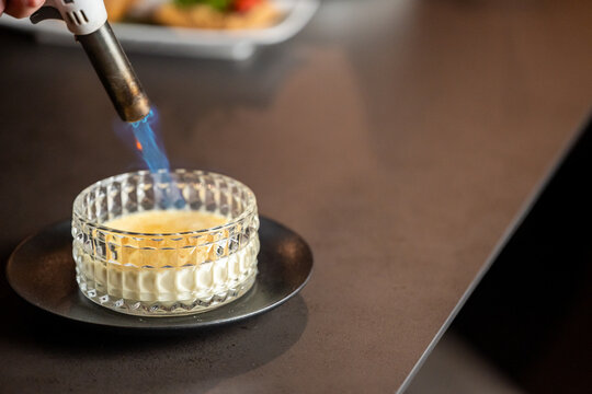 Chef caramelizing crème brûlée with kitchen torch, close-up of dessert in glass ramekin on dark plate, gourmet cooking technique in professional kitchen.