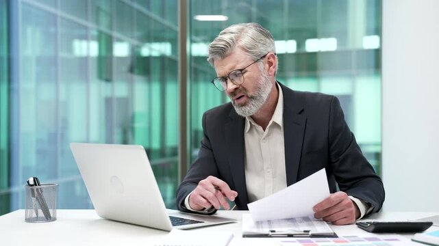 Frustrated mature businessman unhappy with financial results sitting at desk in business office. Upset puzzled financier reviewing documents and looking at laptop screen, unhappy with bad indicators