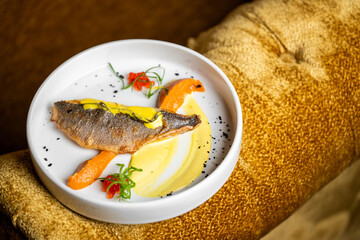 Gourmet fish fillet with yellow sauce, puree, roasted carrot, herbs, and sesame seeds on a white plate. Elegant fine dining presentation on plush golden background