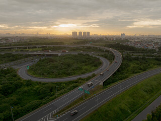 An elevated view of a busy highway interchange in Hanoi at sunset, with cars and trucks navigating the winding ramps and overpasses, all set against a backdrop of a city skyline.