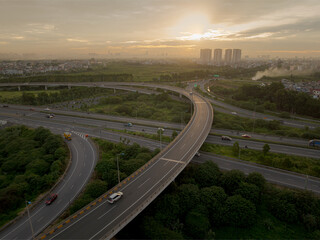 An elevated, wide-angle view of a highway interchange in Hanoi at sunset, showing the sprawling ramps, bridges, and lanes enveloped by lush greenery with distant high-rise buildings on the horizon.