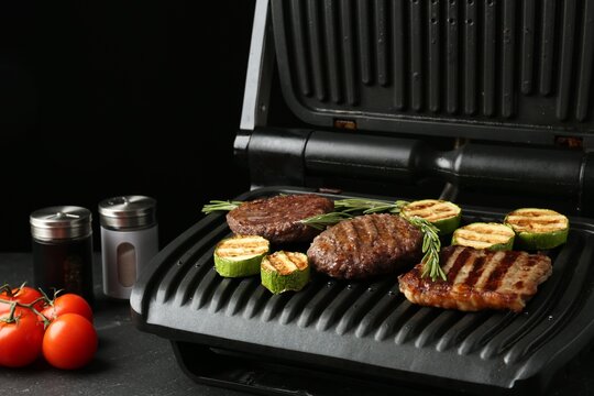Electric grill with beef steaks, zucchini and rosemary on black table, closeup