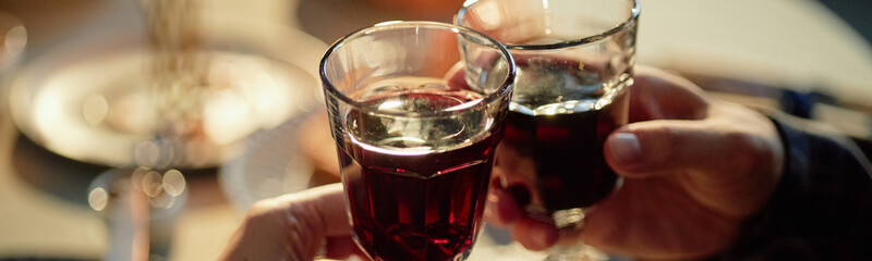 Header of adult hands holding wine glasses clinking together during Hanukkah celebration, blurred festive table setting visible in background, capturing traditional holiday toast moment