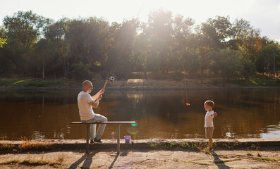 Father and young son fishing at calm lake in countryside at sunset. Horizontal family lifestyle photo with nature background and warm summer mood.