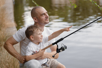 Father and young son fishing at calm lake in countryside at sunset. Horizontal family lifestyle photo with nature background and warm summer mood.