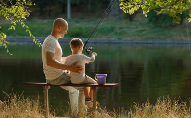 Father and young son fishing at calm lake in countryside at sunset. Horizontal family lifestyle photo with nature background and warm summer mood.