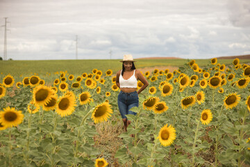 Fototapeta premium Young, attractive, Latina brunette woman wearing a white crochet top, jeans, and a hat, smiling and happy as she walks through a field of sunflowers. Concept: nature, agriculture, countryside.