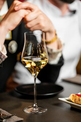 Close-up of a wine glass with white wine on a dark table in a formal dining setting, with blurred people and food in the background. Elegant atmosphere, lifestyle concept.
