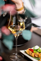Close-up of white wine glass and gourmet dumpling with herbs and sauce on elegant table setting. Blurred background with foliage and hand adds depth and fine dining atmosphere.