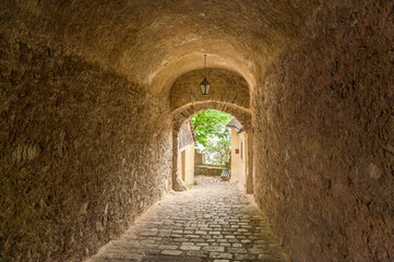 Obraz premium An archway leads into a quaint courtyard in Hallstatt, Austria, featuring vibrant greenery and vintage cobblestones illuminated by soft natural light, creating an inviting atmosphere reminiscent of