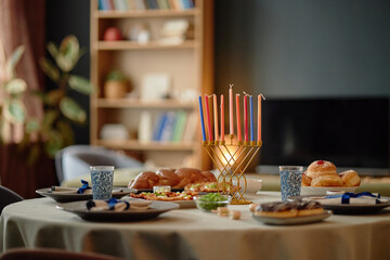 Hanukkah table setting featuring menorah with lit candles standing on round table surrounded by traditional Jewish holiday foods including challah bread, sufganiyot, and potato pancakes
