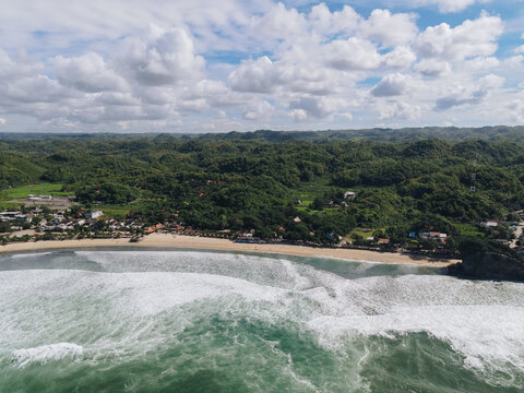 Aerial view of the beach meeting the forest under a vast sky, the waves crash in white foam against the shore, Pacitan, East Java, Indonesia.