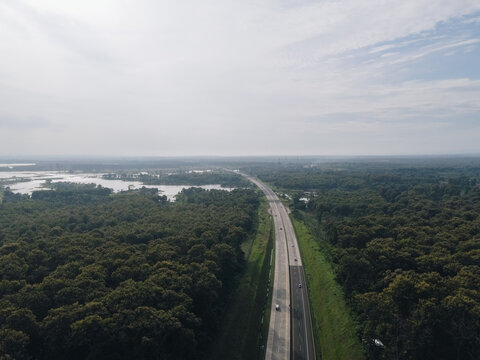 Aerial view of a straight highway cutting through a dense forest, the road ahead disappearing into a hazy horizon, [Actual City Name], [Actual State Name], [Actual Country Name].