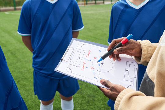 Teenage boys standing on soccer field listening to Black man coach explaining game strategy using clipboard with tactical diagram during team practice session