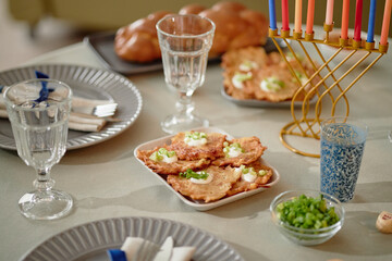 Hanukkah table setting featuring golden brown potato latkes garnished with green onions, traditional menorah with colorful candles, braided challah bread, and napkins arranged for celebration