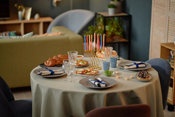 Round table set for Hanukkah celebration featuring menorah with colorful candles, traditional challah bread, latkes, sufganiyot, glassware and festive place settings