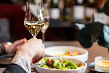 Close-up of a hand holding a glass of white wine at a restaurant table with gourmet food. Warm lighting, elegant atmosphere, fine dining and social gathering
