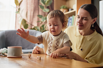 Caucasian young mother watching toddler playing with wooden dreidels on table during Hanukkah celebration, both sitting together in cozy home setting, mother smiling gently