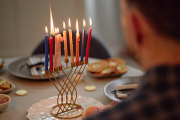 Caucasian man sitting at table celebrating Hanukkah, watching lit menorah candles burning,...