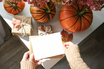 Cozy autumn flat lay with hands in sweater holding blank greeting card, surrounded by pumpkins, wrapped gifts and fall leaves on white surface. Ideal for seasonal greetings and Thanksgiving