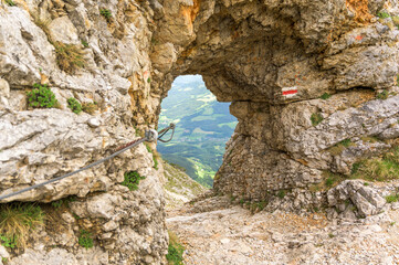 A stunning natural rock arch frames a breathtaking view of the lush green valley and hills of Rax Austria, with sunlight casting a warm glow over the landscape and vibrant blue skies