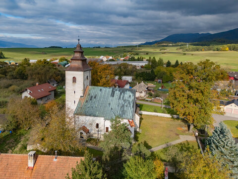 Aerial view of the stone church with a dark steeple stands amidst a village, framed by golden trees and distant mountains, H&Atilde;&iexcl;j, &Aring;&frac12;ilina Region, Slovakia.