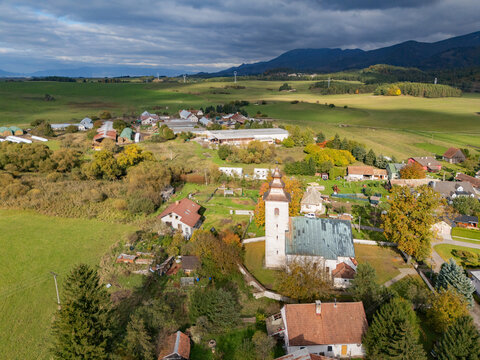 Aerial view of a quaint village nestled among rolling green hills with a striking white church as its centerpiece, under a dramatic sky, H&Atilde;&iexcl;j, &Aring;&frac12;ilina Region, Slovakia.
