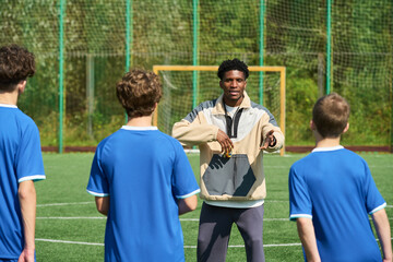 Black man coaching group of teenage boys on outdoor soccer field, gesturing with hands while giving instructions, goalpost and netting in background