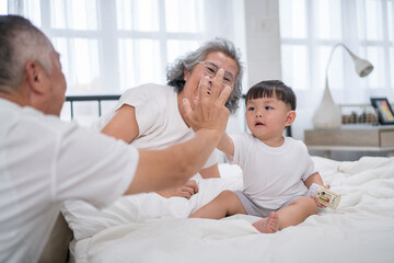 Happy Asian grandparents bonding with toddler grandson on bed, smiling and giving high five in cozy bedroom. Concept of intergenerational love, grandparenting, and joyful family moments at home.