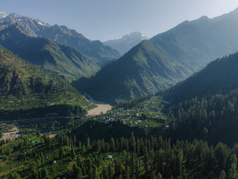 Aerial view of a small village nestled in a valley with a river flowing through it, surrounded by towering green mountains and snow-capped peaks, Tosh, Himachal Pradesh, India.