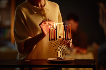 Woman lighting candles on menorah during Hanukkah celebration, holding lit candle with both hands, blurred figure of man sitting in background