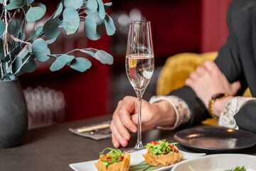 Elegant dining scene with man in suit holding champagne, gourmet appetizers on white plate, black tableware, and eucalyptus centerpiece in upscale restaurant setting