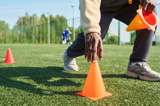Black man crouching on sports field placing orange training cone on artificial grass, holding additional cones in other hand, wearing athletic shoes, soccer players visible in background - Powered by Adobe