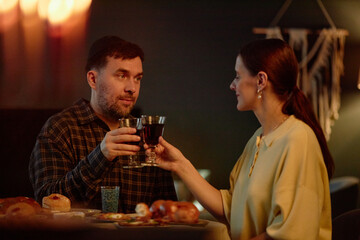 Caucasian middle aged man wearing kippah and woman clinking wine glasses while sitting at table with traditional Hanukkah foods and menorah in background