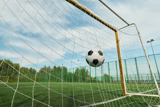 Soccer ball hitting net inside outdoor goal on green field, capturing moment of scoring during game, metal goalposts and tall fence surrounding pitch under blue sky