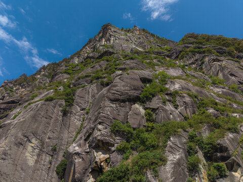 Aerial view of the rugged, textured mountain face, etched with shadows and dotted with resilient green vegetation under a bright sky, Sethan, Himachal Pradesh, India.