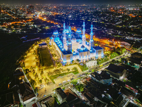 Aerial view of Sheikh Zayed Grand Mosque illuminated in ethereal blue under the night sky, contrasting with the warm glow of the surrounding city lights, Surakarta, Central Java, Indonesia.