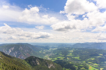 Naklejka premium The image showcases a stunning panoramic view of Rax Mountains in Austria under a clear blue sky with fluffy white clouds, featuring vibrant green hillsides leading into a lush vall