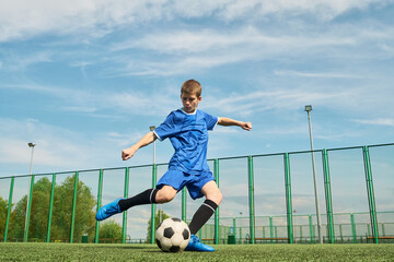 Teenage boy kicking soccer ball on outdoor field, wearing sports uniform, showing athletic movement...
