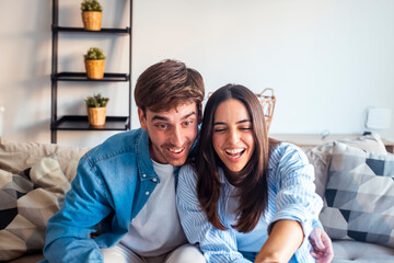 Modern couple sitting on the sofa at home using a laptop together, smiling and laughing during...