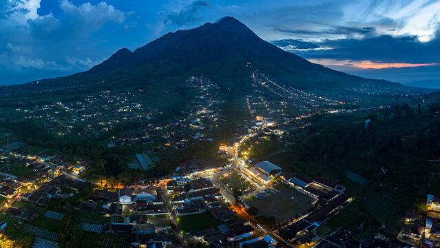 Aerial view of the majestic Mount Merapi looms over a town illuminated with twinkling lights, casting long shadows as dusk settles, Boyolali, Central Java, Indonesia.