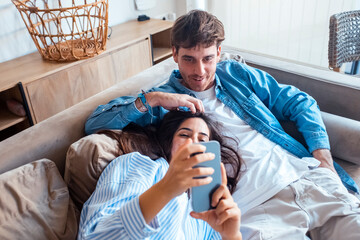 Young couple relaxing at home on sofa, woman lying with head on man’s lap while he uses...