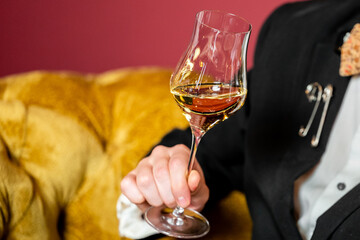 Stylish man in formal suit with bow tie and safety pins on lapel, holding glass of amber liquor, seated on mustard-yellow tufted couch against red background.