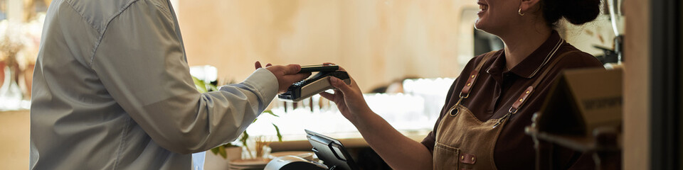 Caucasian man handing payment terminal to smiling young adult Caucasian woman working behind...