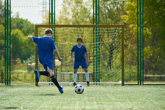 Teenage boy kicking soccer ball toward goal while another teenage boy standing as goalkeeper on outdoor field during daytime sports practice session